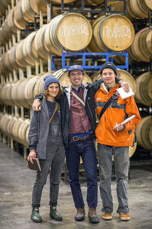 Three people stand together in front of a wall of stacked wooden barrels. They are dressed in casual outdoor clothing, with coats, hats, and boots. They are smiling and have their arms around each other, suggesting camaraderie. The barrels have markings on them.