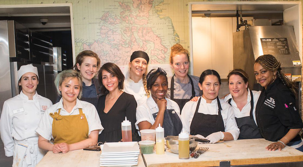 A group of ten smiling women wearing various chef uniforms and casual clothing stand behind a counter in a kitchen. Several kitchen items, including sauce bottles and a stack of plates, are on the counter. A map is visible on the wall behind them.