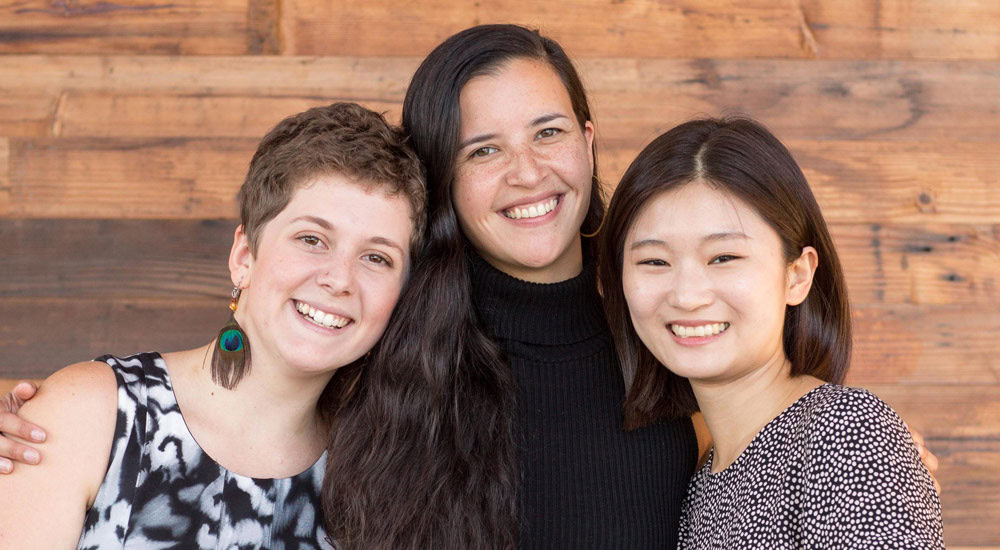 Three smiling people stand close together in front of a wooden wall. The first person on the left has short, curly hair, the person in the middle has long, straight hair, and the person on the right has straight, shoulder-length hair. They appear to be happy.