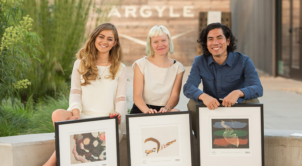 Three people are sitting outside, each holding a framed piece of artwork. They are smiling and posing for the photo. The background includes greenery and a building with the word 