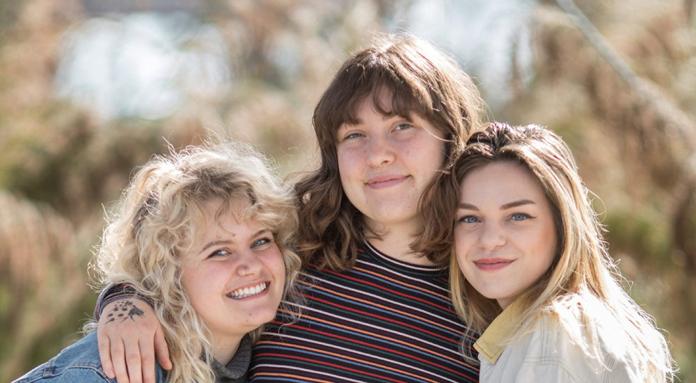 Three people are smiling and standing close together outdoors. The person on the left has curly blond hair, the person in the middle has wavy brown hair, and the person on the right has straight blond hair. They appear happy and it is a sunny day.