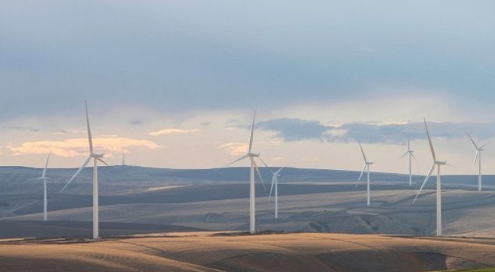 Wide landscape view of a wind farm with several wind turbines spread across rolling hills. The sky is cloudy with patches of soft sunlight breaking through, illuminating parts of the serene landscape.