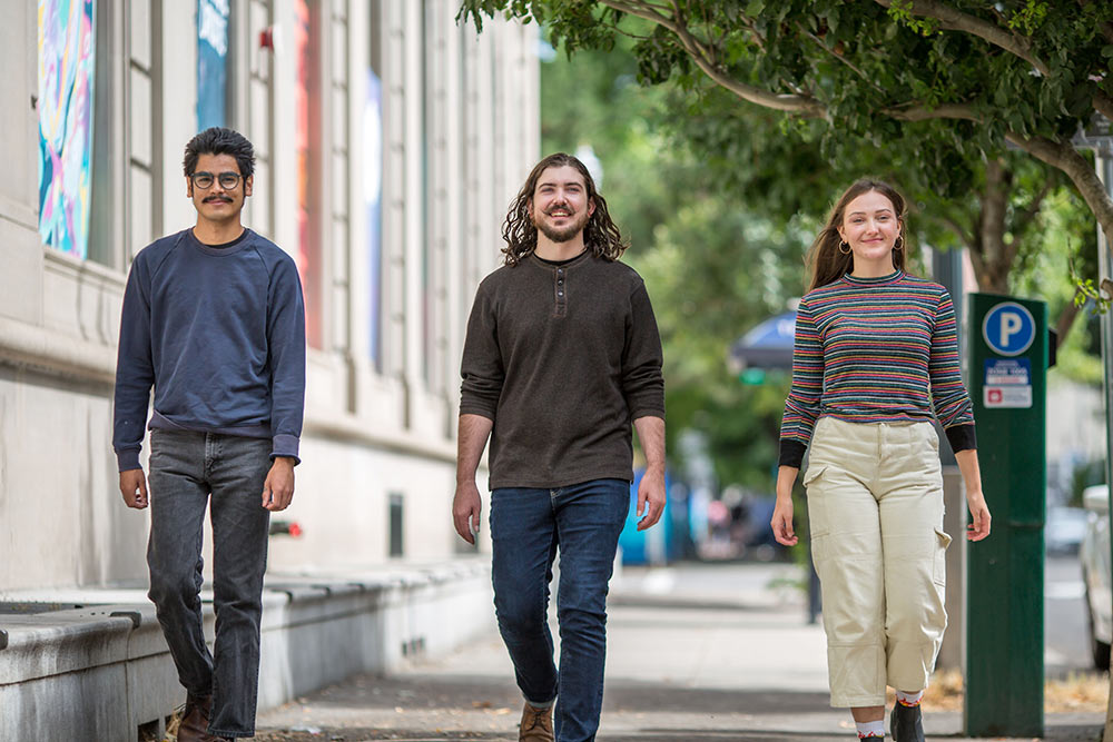 Three people are walking down a sidewalk on a sunny day. The group consists of a man with glasses and a mustache, a man with long hair and a beard, and a woman with long hair. They are smiling, dressed casually, and appear relaxed. Trees line the path.