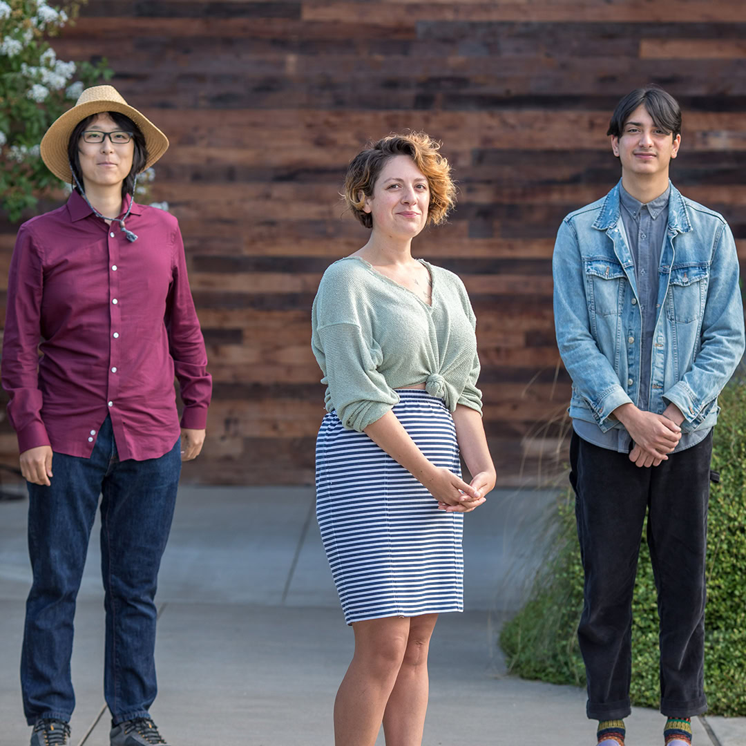 Three people stand outside on a paved area in front of a wooden wall. The first person on the left wears a sunhat, glasses, a maroon shirt, and jeans. The middle person wears a light green top and a striped skirt. The third person wears a denim jacket and black pants.