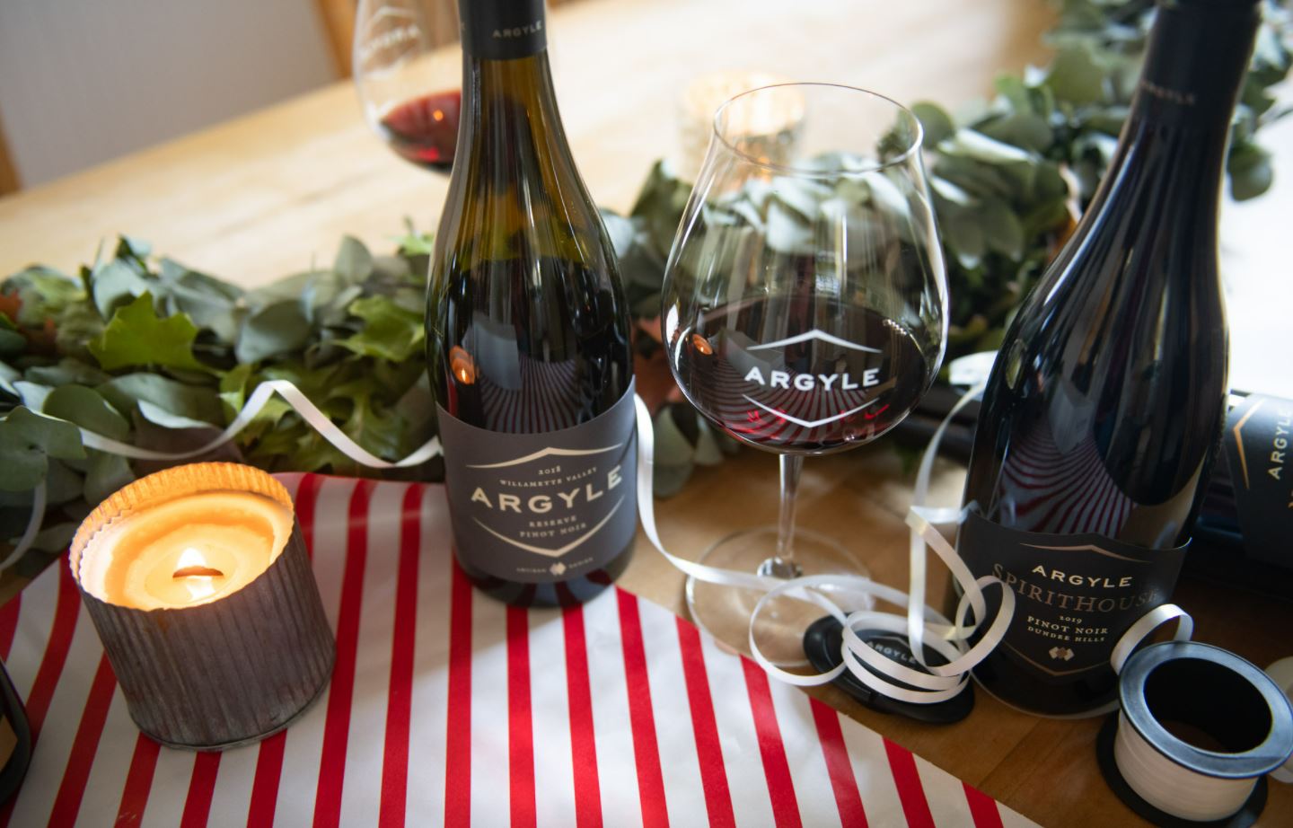 A festive table setup features two Argyle wine bottles, a glass of red wine, a lit candle, a green leafy garland, and decorative ribbons. The striped red and white tablecloth adds to the celebratory ambiance.