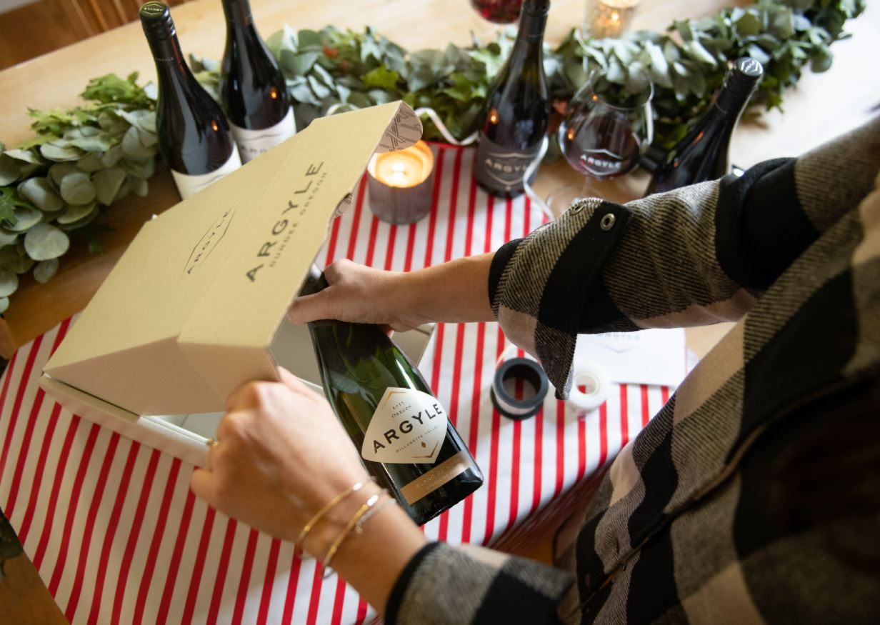 A person wearing a plaid shirt is placing a bottle of Argyle wine into a white gift box on a table. The table is decorated with a red-striped tablecloth, eucalyptus leaves, candles, and other wine bottles and glasses.