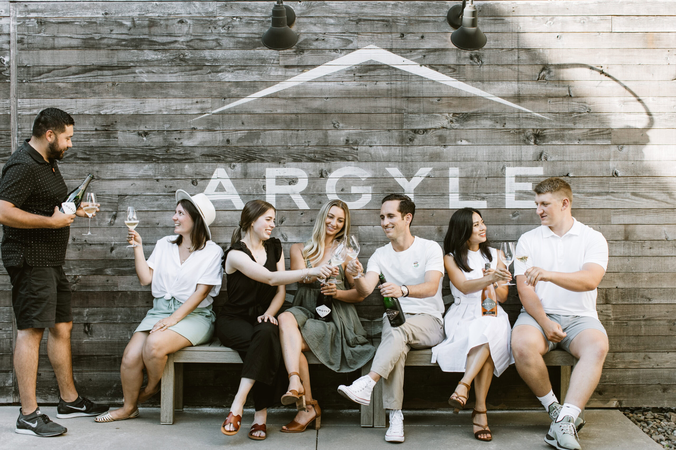 A group of six people sits on a bench against a wooden wall with the word Argyle embossed on it. They are smiling and holding wine glasses, toasting together. One person stands on the left, pouring wine. The setting appears relaxed and celebratory.