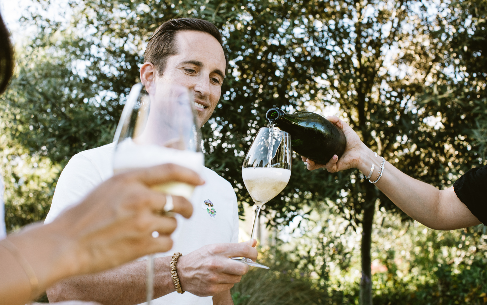 A person smiles while holding a wine glass, which is being filled with champagne by another person. Another hand holding a champagne glass is visible in the foreground. They are outdoors with greenery in the background.