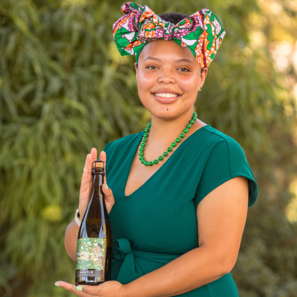 A woman in a green dress and vibrant patterned headwrap holds a wine bottle. She has a green beaded necklace and smiles while posing outdoors with greenery in the background.