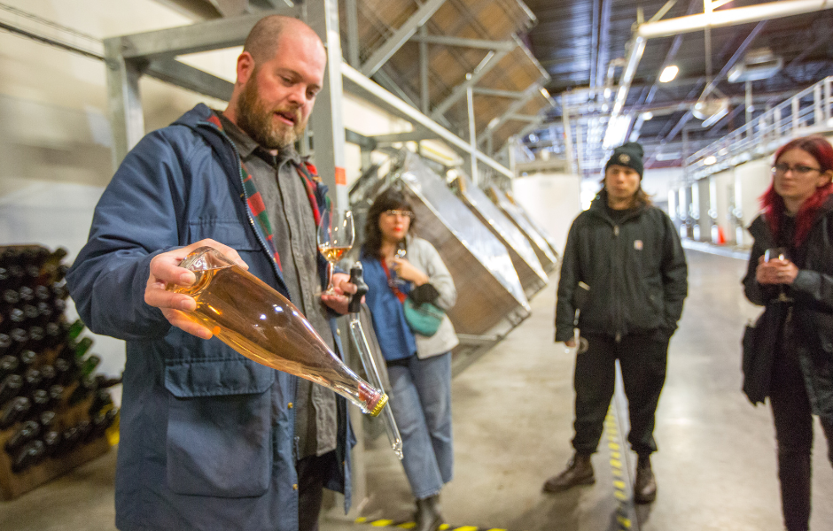 A man in a blue jacket is demonstrating a wine bottle with people watching in an industrial setting. He is tilting the bottle to let some liquid pour out. Others are observing closely, and a woman in a denim jacket is holding a wine glass.