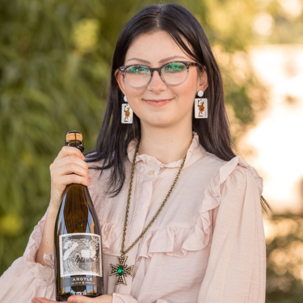 A person with long dark hair, wearing glasses, a pale pink ruffled blouse, and decorative earrings, stands outdoors holding a bottle of Argyle wine with a smile. The background is blurred greenery.