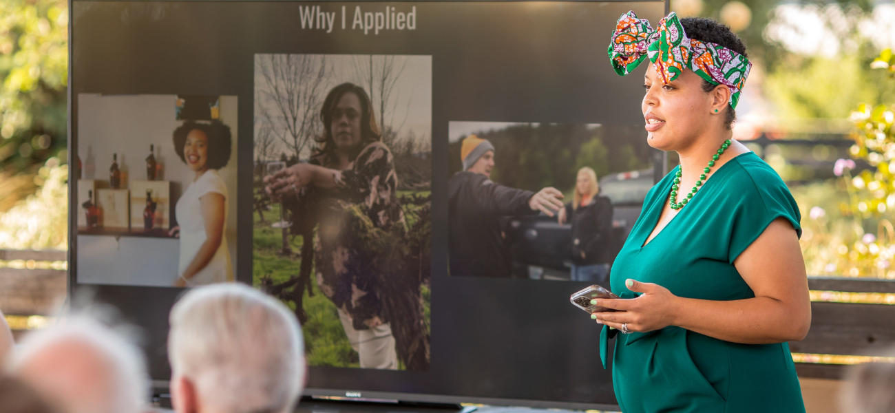 A woman in a green dress and colorful headwrap is presenting outdoors to a small audience. Behind her is a large screen displaying three images of herself and the title 