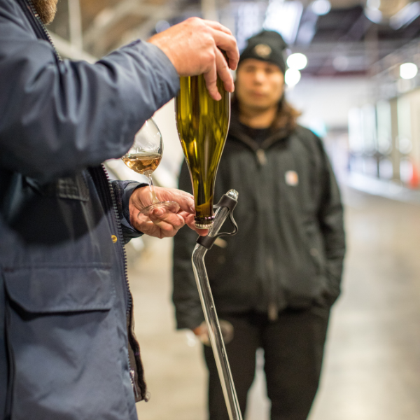 A person in the foreground holds an upside-down brown wine bottle, pouring the liquid into a wine glass. Another person in the background looks on, wearing a black jacket and a beanie, with an industrial setting visible around them.