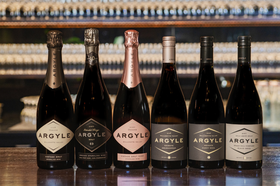 Six bottles of Argyle wine are lined up on a bar counter. The labels indicate a variety of types, including sparkling wine and Pinot Noir. The background is blurred but shows rows of glasses and a dark wooden shelf, creating a sophisticated ambiance.