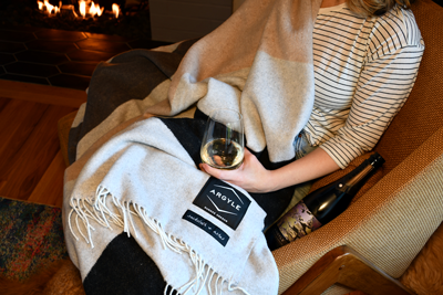 A person sits in a cozy chair by a fire, covered with a large, checkered Argyle blanket. They hold a glass of white wine, with an unopened bottle beside them. Warm lighting enhances the relaxed, cozy ambiance of the scene.