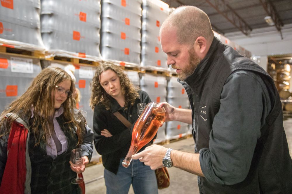 A man holding a bottle of rosé demonstrates it to two women in a warehouse with stacked metal containers. The women watch attentively as the man explains, pointing to the bottle. The scene suggests a wine tasting or educational tour.