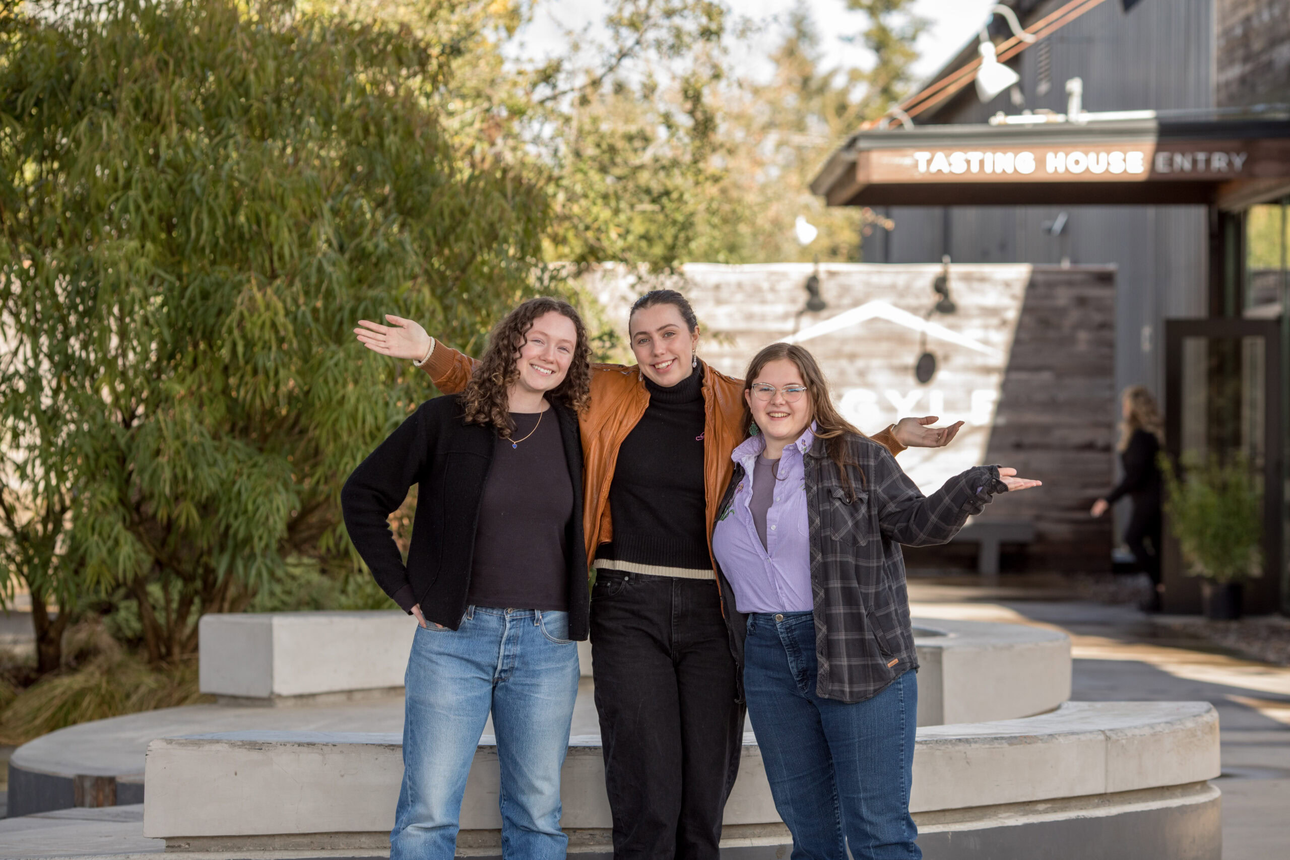 Three people stand outdoors, smiling with arms outstretched. They are posed in front of a tasting house with trees and greenery in the background. The individual on the left wears a black jacket, the one in the middle an orange jacket, and the one on the right a plaid shirt.