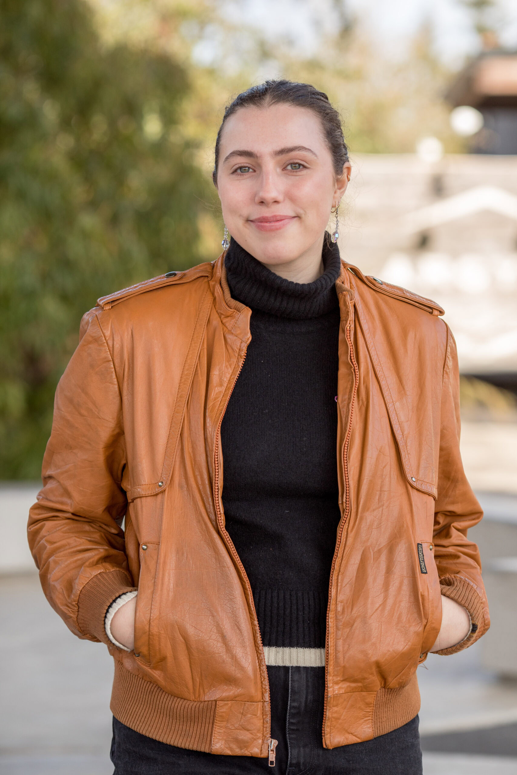 A woman with dark hair pulled back is wearing a brown leather jacket over a black turtleneck sweater. She has her hands in her jacket pockets and is smiling slightly. The background shows an outdoor setting with blurred greenery and structures.