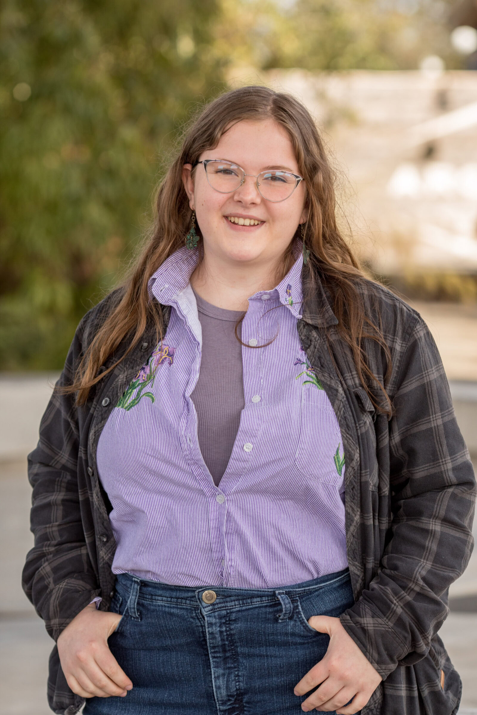 A person with long hair and glasses smiles while standing outdoors. They are dressed in a striped shirt with floral embroidery, a dark plaid jacket, and jeans. The background includes greenery and blurred outdoor elements.