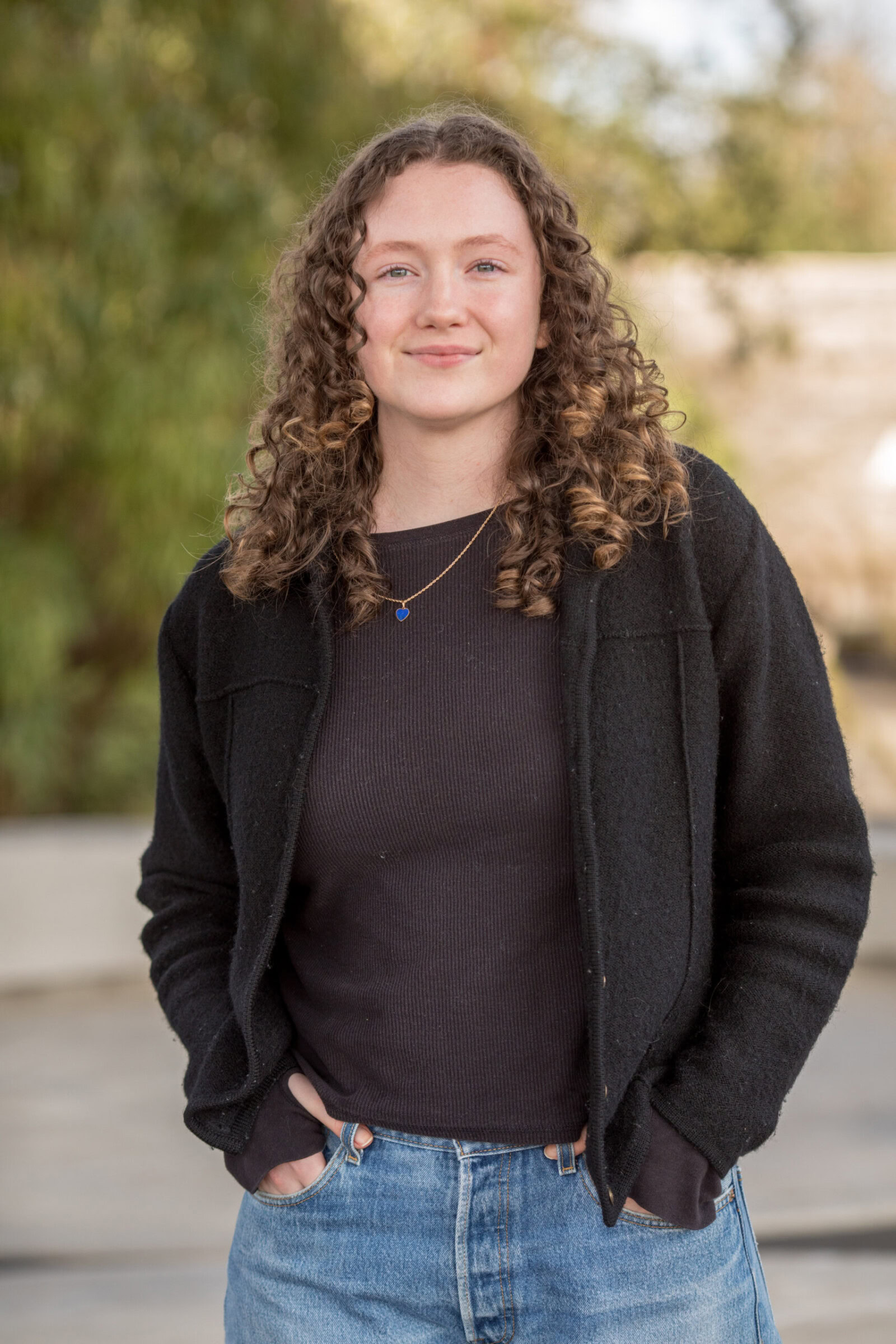 A young person with curly hair stands outdoors, facing the camera and smiling slightly. They are wearing a black jacket over a dark top and light blue jeans, with hands in their pockets. The background features blurred greenery and a light-colored structure.