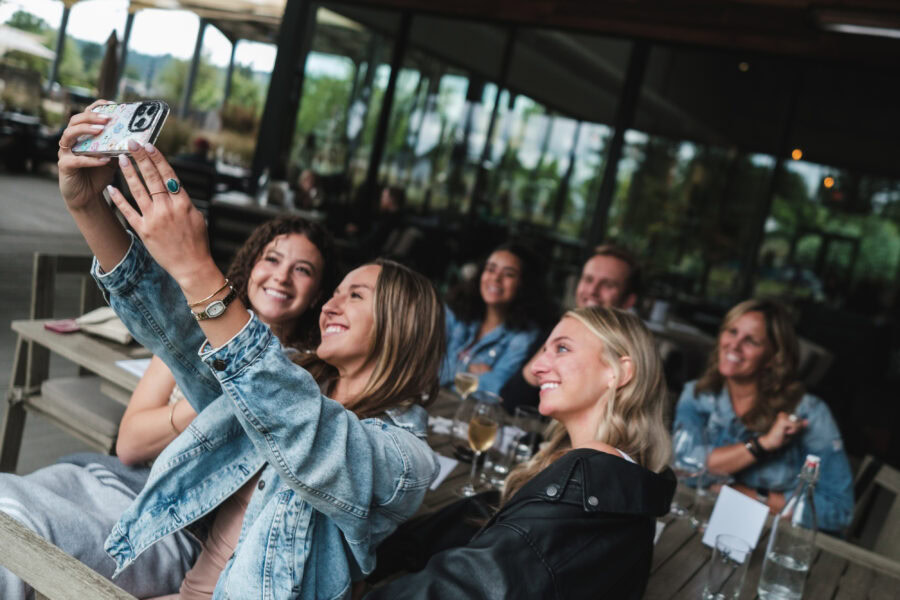 A group of six friends are sitting at the Argyle Tasting House and smiling as two women in the front take a selfie with a smartphone. They are surrounded by wine and appear to be enjoying a casual gathering.