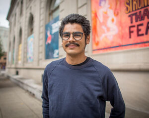 A person with dark hair, glasses, and a mustache is smiling while standing outdoors in front of a building with colorful murals on the wall. They are wearing a dark long-sleeve shirt and behind them, the sidewalk extends along the length of the building.