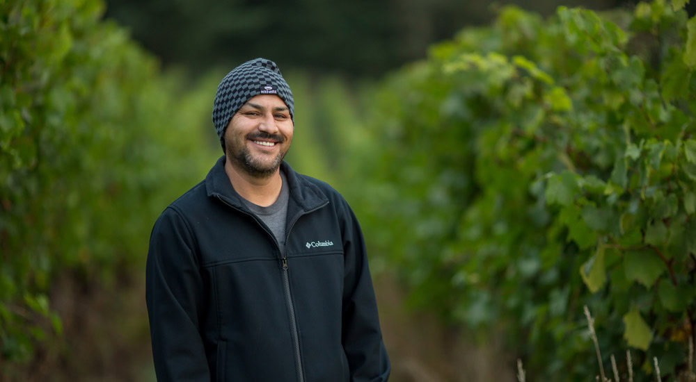 A person wearing a black Columbia jacket and a knitted hat smiles while standing in a lush vineyard with rows of green grapevines extending into the background. The individual appears relaxed and happy.