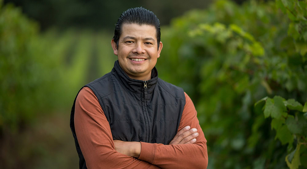 A person with dark hair, wearing a black vest over an orange long-sleeve shirt, stands outdoors with arms crossed and a smile on their face. The background features green, leafy bushes in a natural setting.