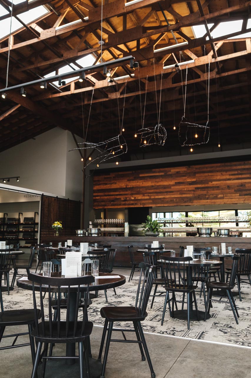 A spacious, modern restaurant with an open ceiling featuring wooden beams. The dining area has black wooden chairs and round tables, each set with glasses and menus. Warm lighting hangs above and the back wall consists of wooden panels, shelves, and a bar area.