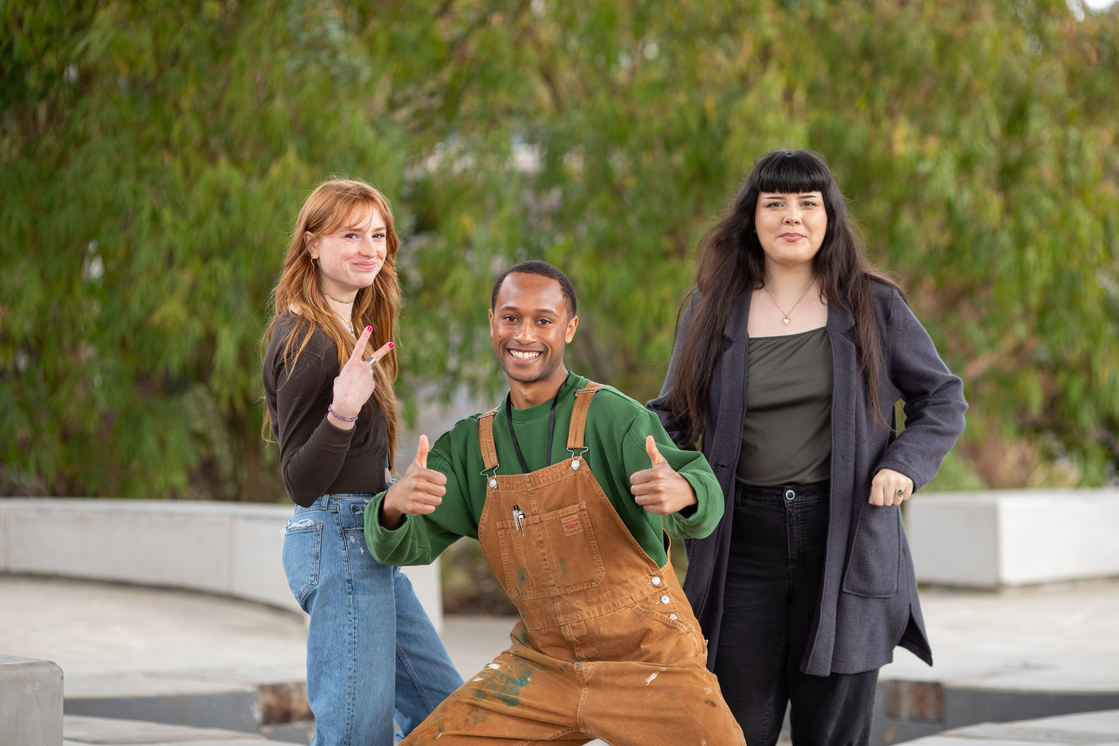 Three people are posing outdoors in front of lush greenery. One person on the left, with long red hair, is showing a peace sign. The person in the middle, wearing brown overalls, is giving two thumbs up. The person on the right, with long black hair, is smiling.