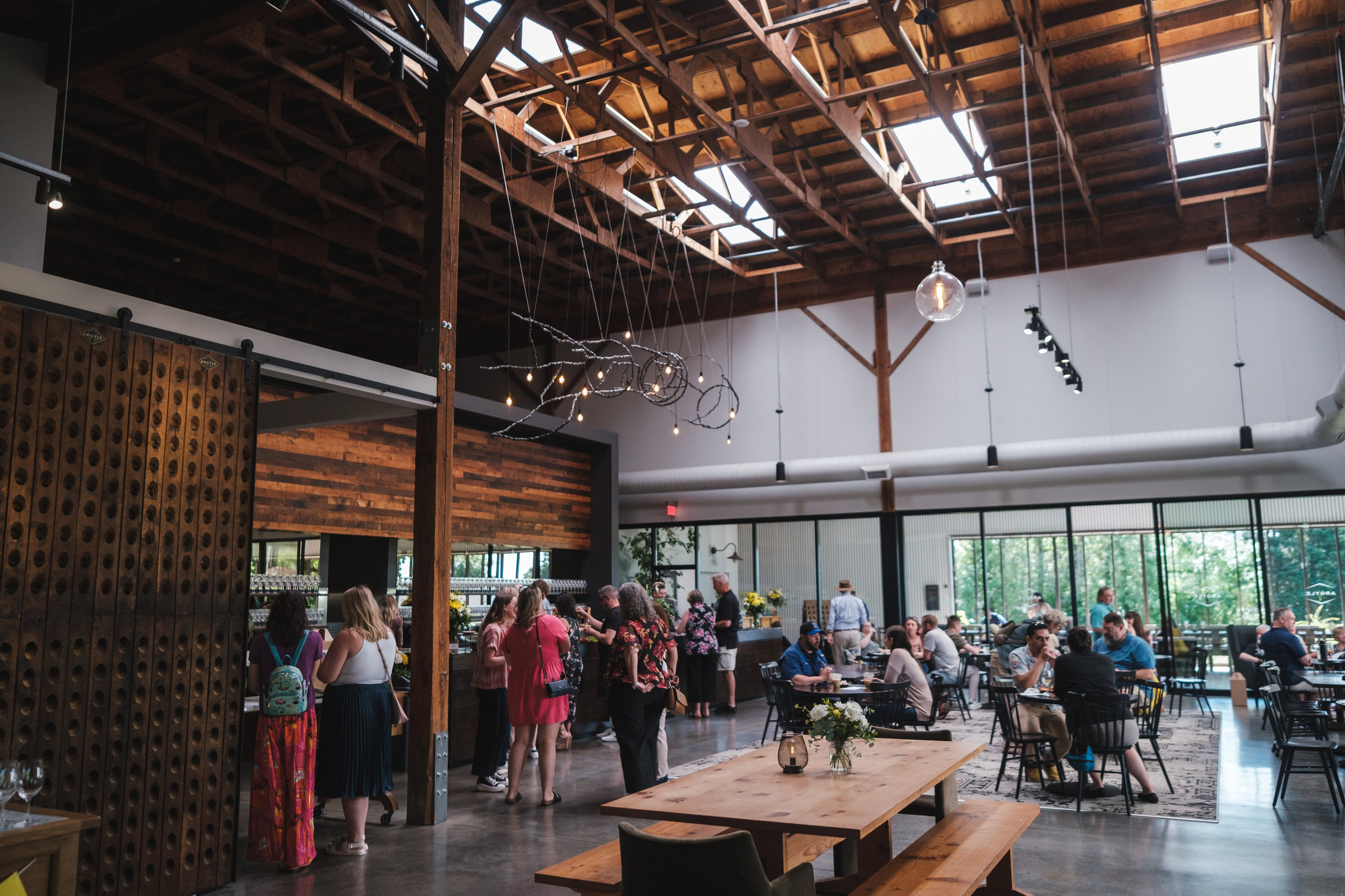 A spacious, modern cafe with high wooden ceilings and large windows. People are gathered around tables, chatting, and enjoying their time. Some are lined up at the counter. The room is decorated with rustic wooden elements and hanging light fixtures.