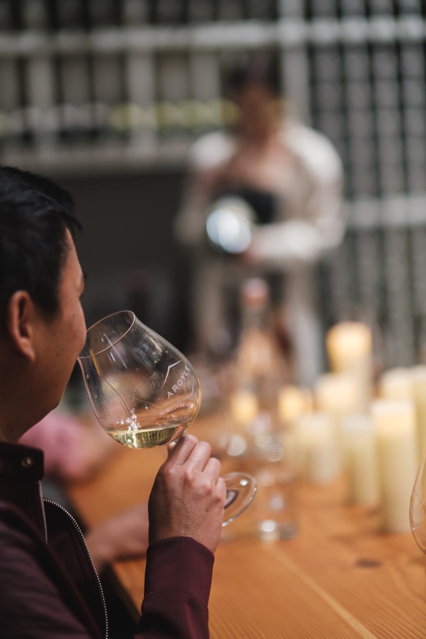 A man holds a wine glass and appears to be sniffing the wine. He is seated at a wooden table adorned with numerous lit candles. In the blurred background, another person is present, possibly engaged in wine service. Wine bottles are visible on shelves behind them.