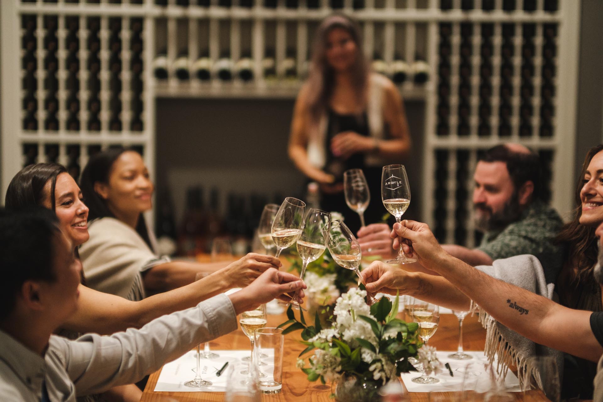 A group of people sitting around a table in a wine cellar, raising their glasses in a toast. Bottles of wine are visible on the shelves behind them. They seem to be enjoying a celebratory gathering. Flowers and wine glasses are on the table.