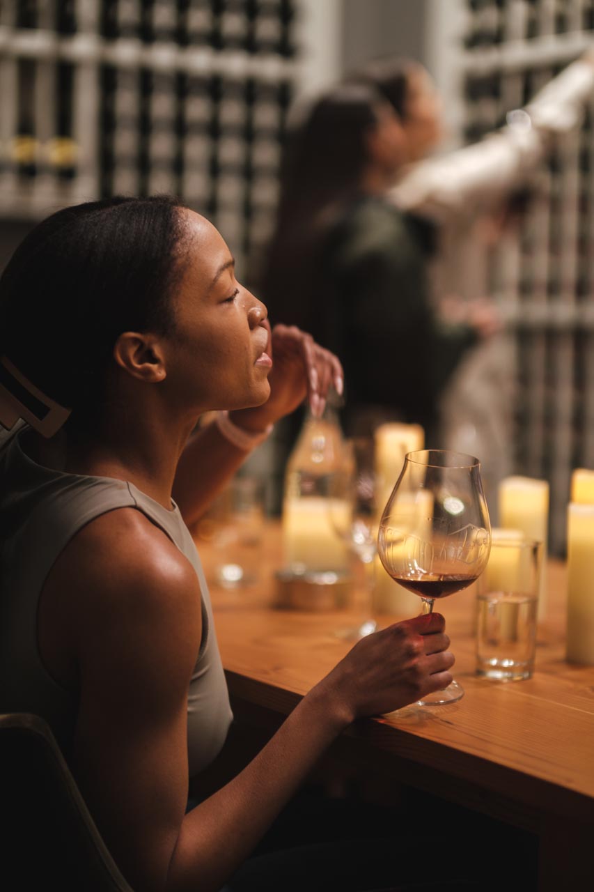 A woman sits at a wooden table holding a glass of wine, immersed in thought. Several candles are lit on the table, creating a warm ambiance. In the blurred background, two other individuals inspect the contents of shelves filled with wine bottles.