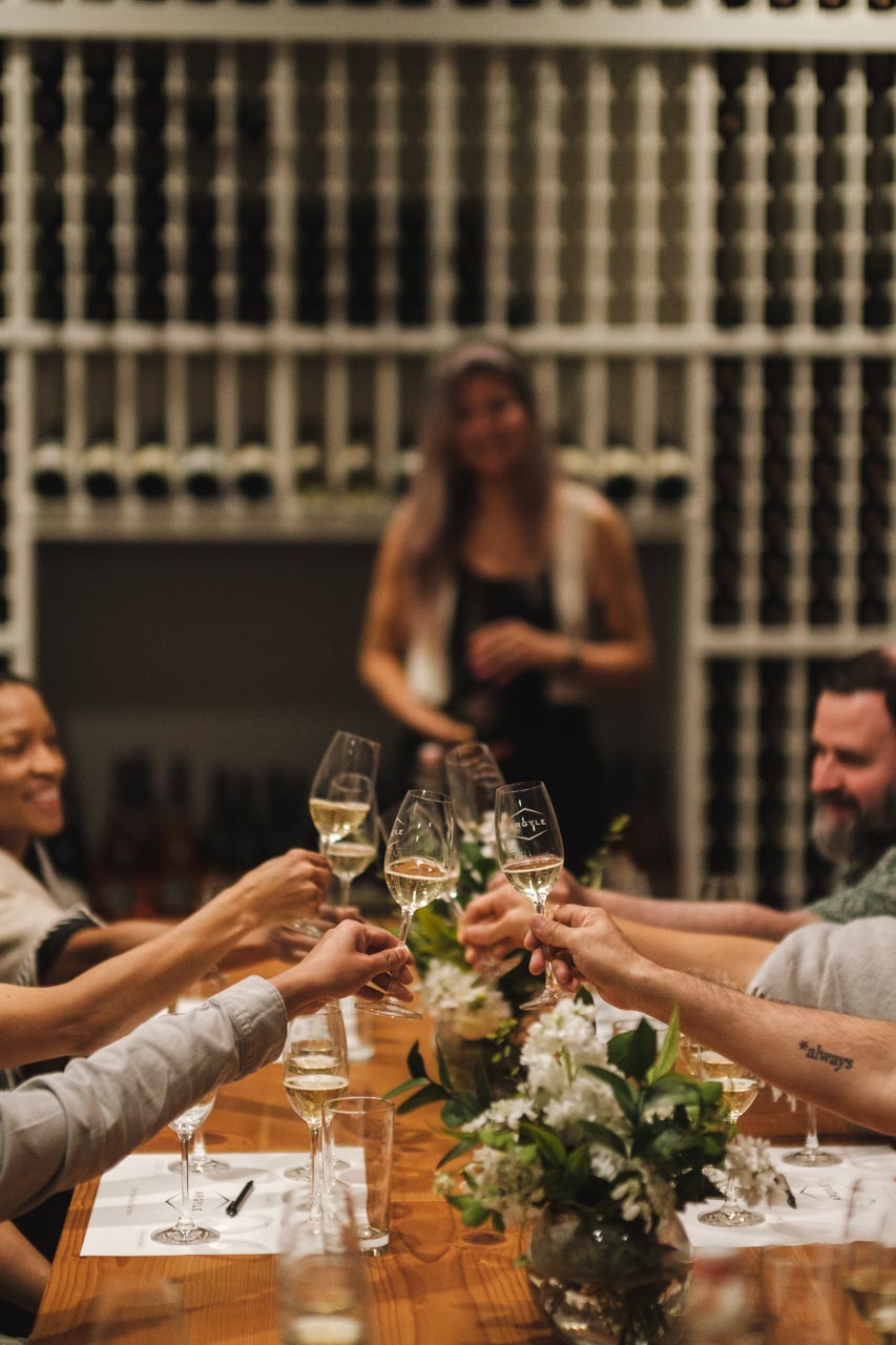 A group of people seated around a wooden table raising their glasses in a toast, with a wine rack-filled wall in the background. A woman stands in the background, holding a wine glass and smiling. The table is decorated with small floral arrangements.