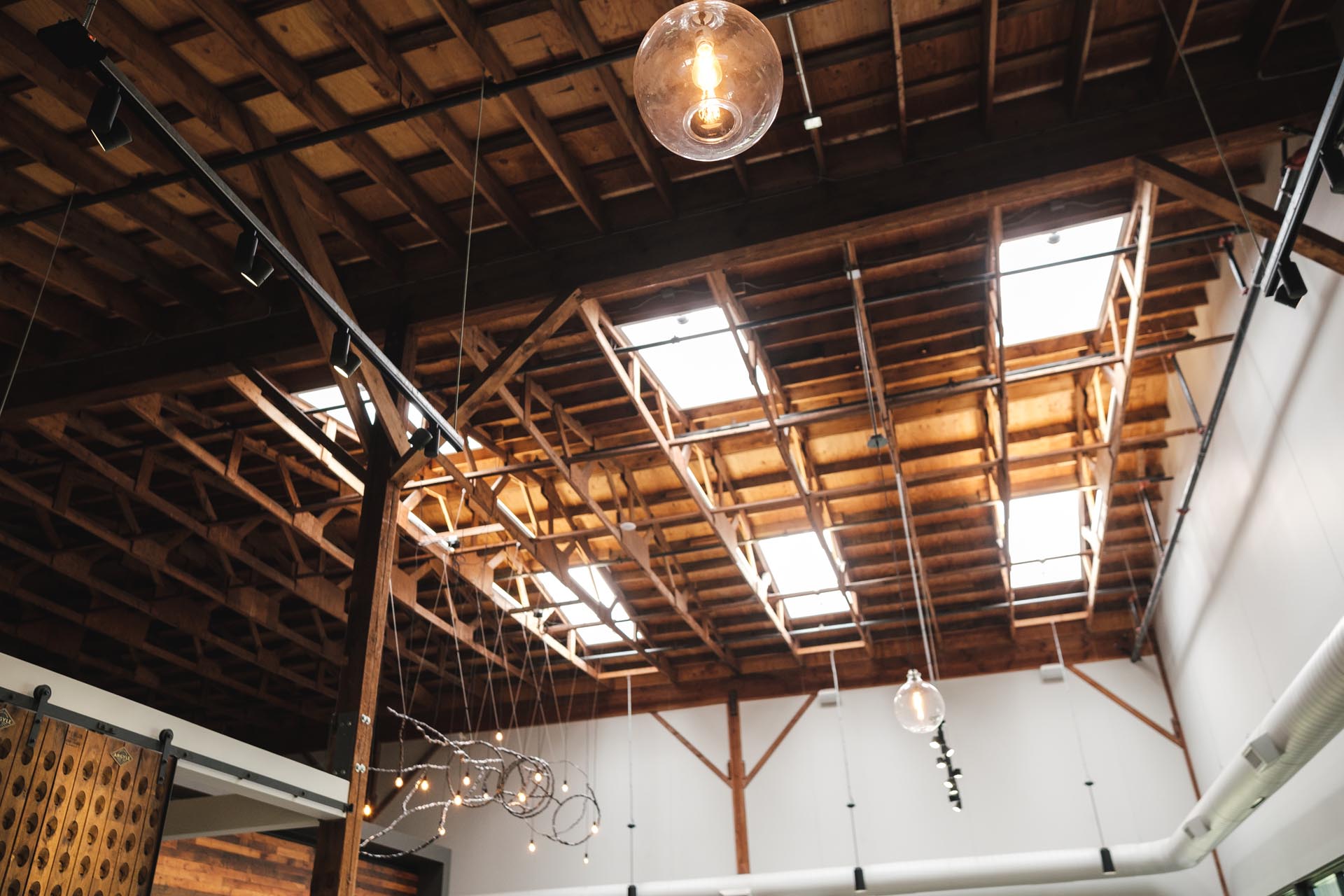 Interior view of a modern building featuring an open wooden ceiling with exposed beams and skylights, reminiscent of Argyle Winery. Several pendant lights and track lighting fixtures hang from the ceiling, creating a rustic yet contemporary ambiance.