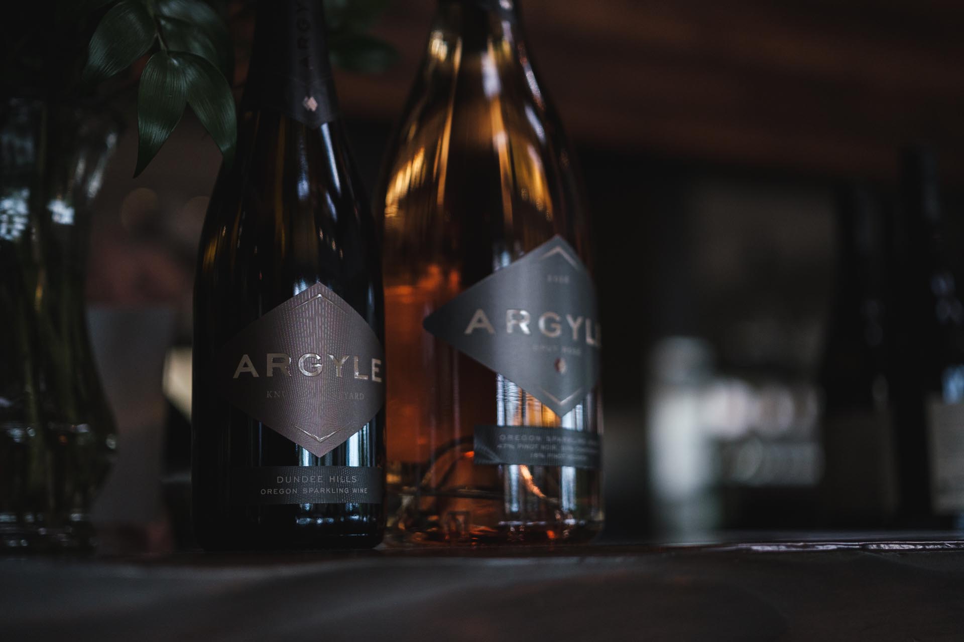 Two Argyle wine bottles, one with a dark label and the other with a gold label, are displayed on a dimly lit surface. Both have 