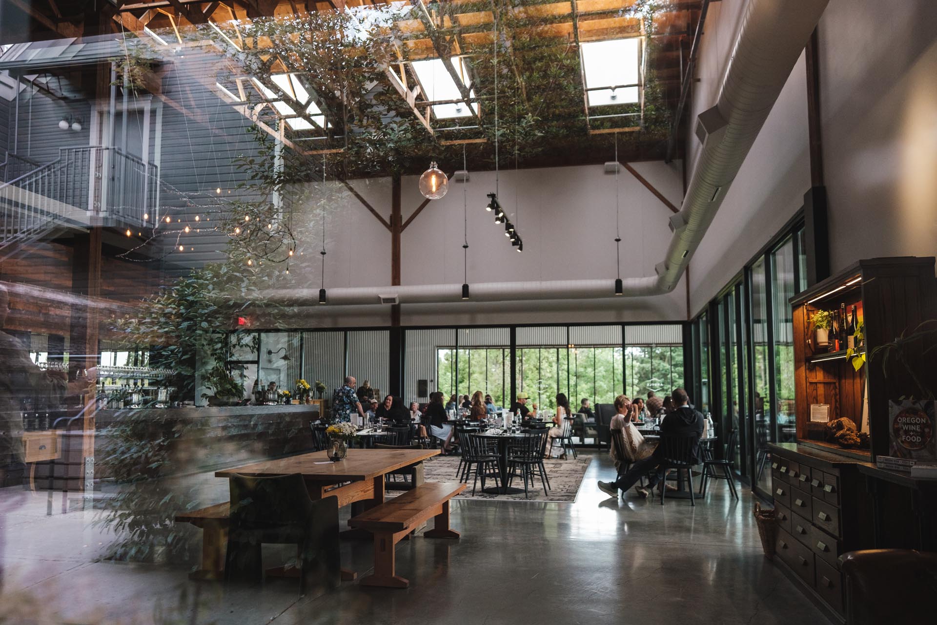 The spacious, modern ARgyle Winery Tasting Room with high ceilings and large windows allowing natural light to flood in. Guests are seated at tables, engaged in conversation. The interior features a mix of wooden furniture, indoor plants, and industrial-style design elements.