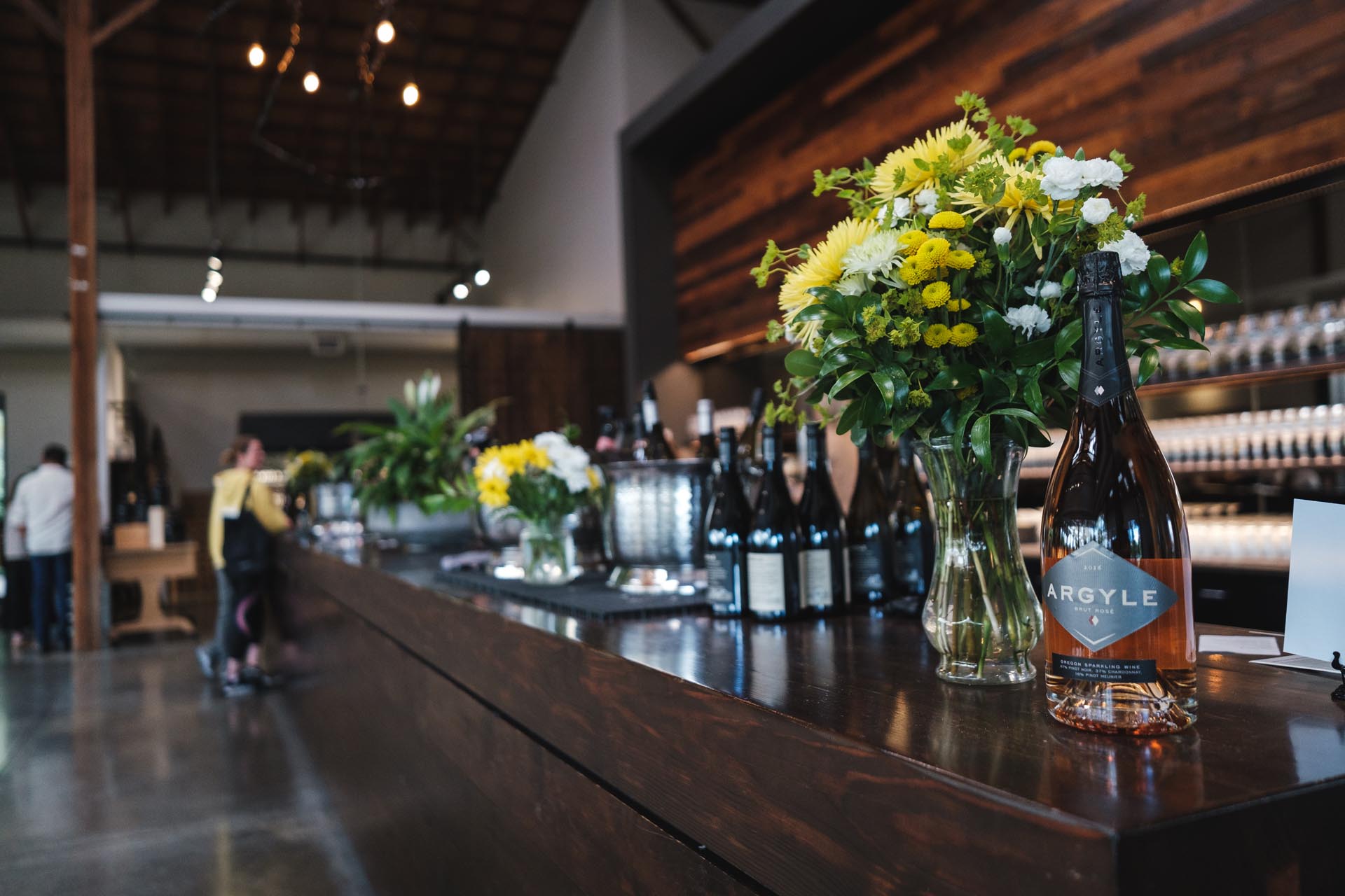 An elegant wine bar with a wooden counter adorned with flower arrangements and a variety of wine bottles. A few patrons are seen in the background, engaging in conversation and exploring the venue's offerings. The space features high ceilings and industrial lighting.