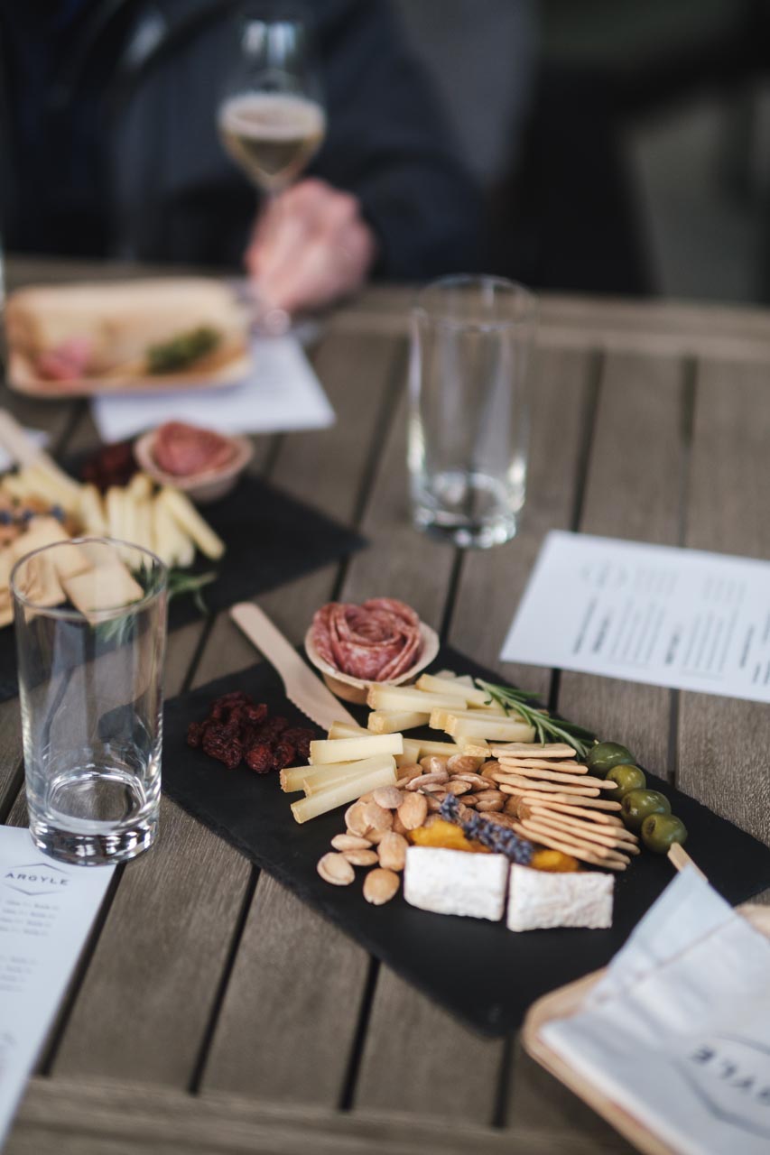 A wooden table with a black slate serving board displaying various cheese assortments, nuts, crackers, cured meats, and green olives. Two empty glasses and a partially visible person holding a beverage are in the background alongside menus on the table.