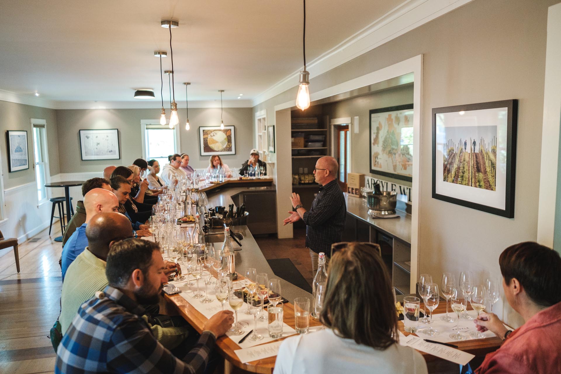 A group of people sit at a long bar counter in a well-lit room with contemporary decor, participating in a wine tasting event. A person stands at the front, speaking, with various wine glasses and bottles arranged on the counter before the seated attendees.