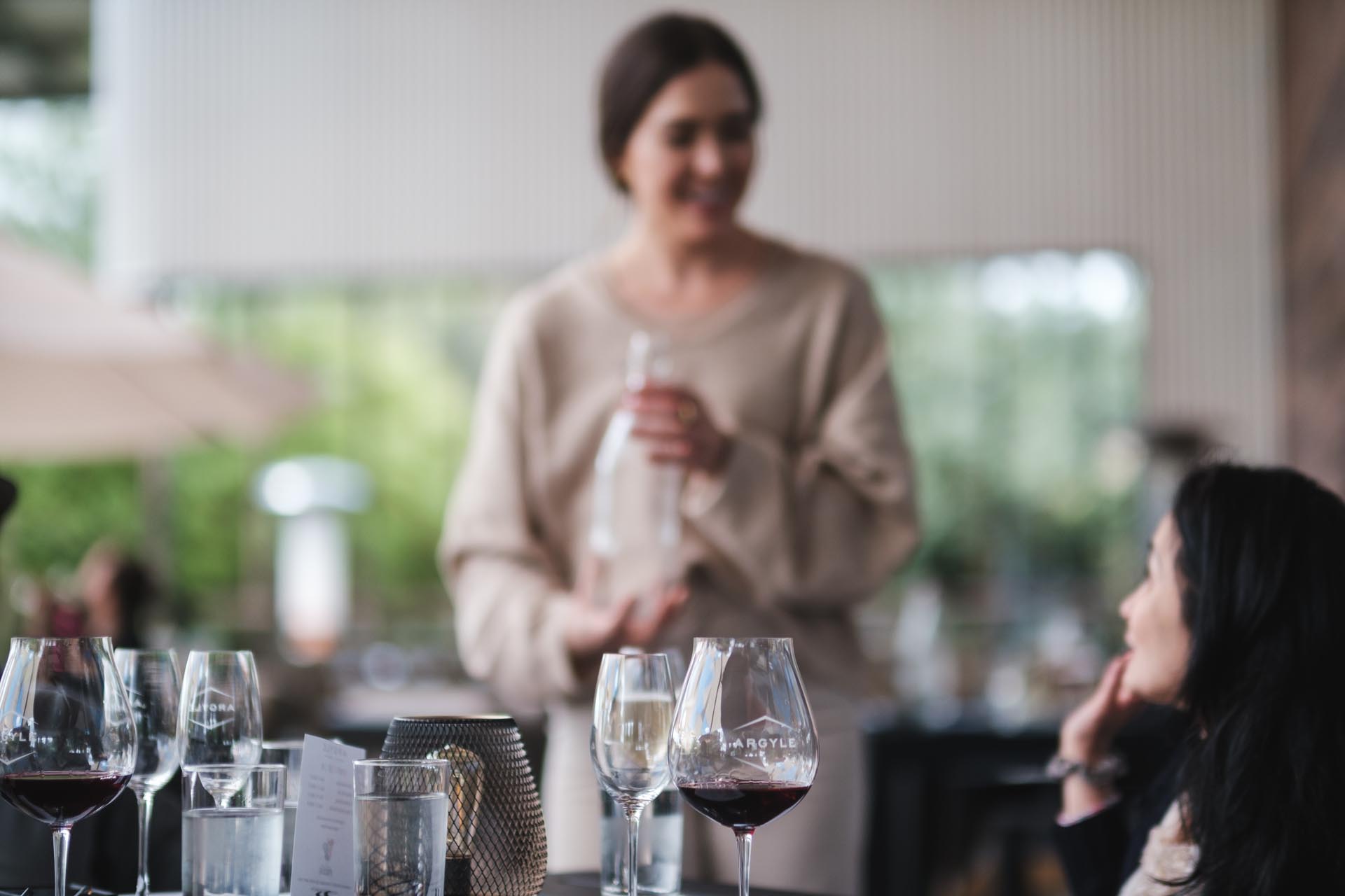 Two women are sitting at a table set with various glasses, including wine and water glasses, and a candleholder. One woman is seated and looking up, while the other stands, seemingly engaged in conversation, holding a bottle from Argyle Winery. The background is blurred and outdoor.