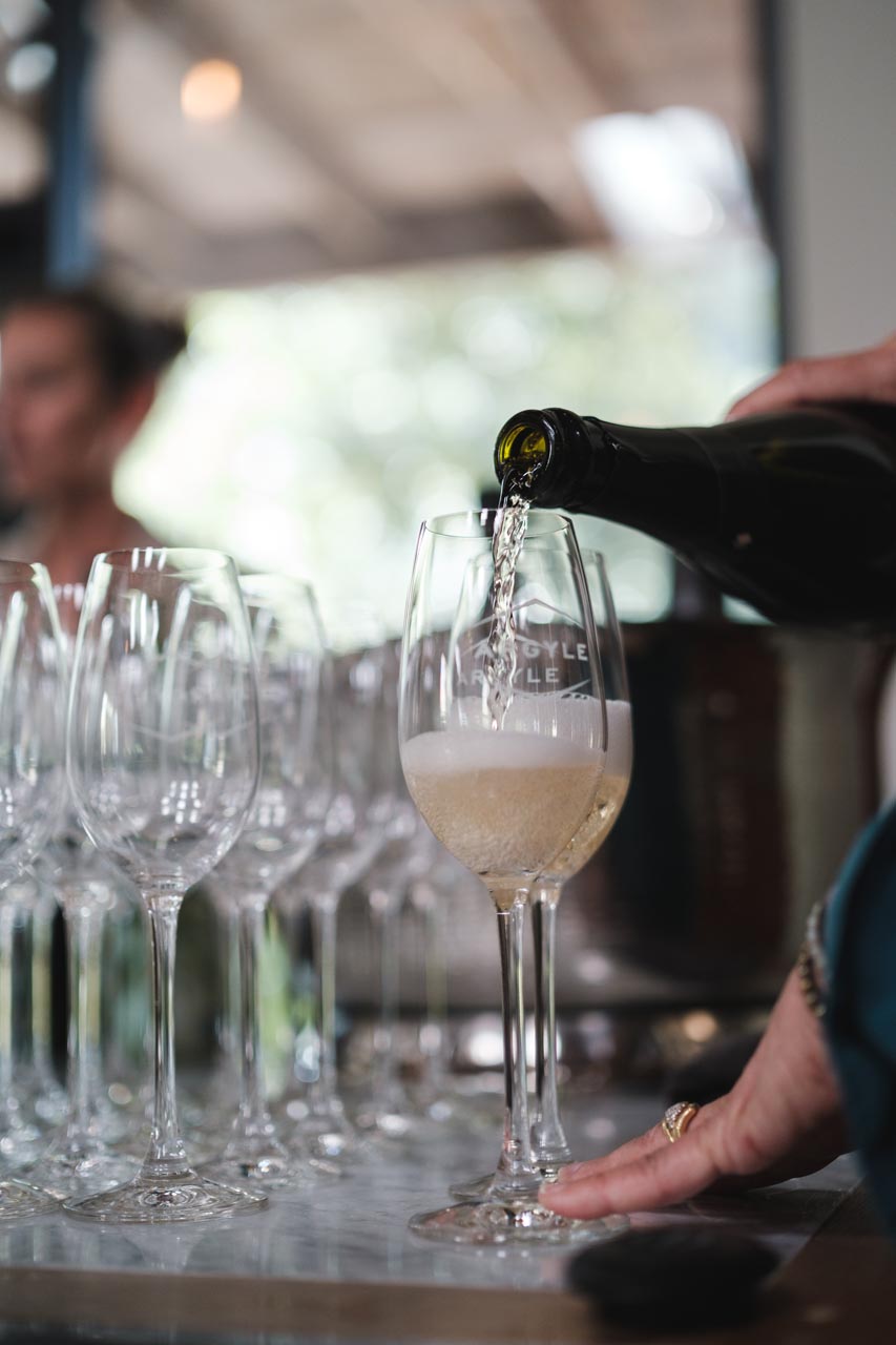 A person pours sparkling wine into a tall, elegant glass. Several empty flutes are lined up next to the filled one on a reflective surface. The background is slightly blurred, focusing on the pouring action and the glass being filled with bubbly liquid.