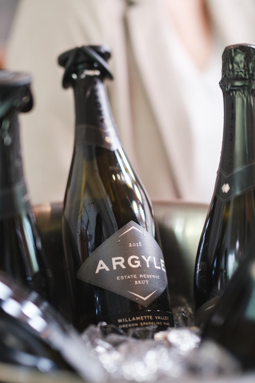 Close-up of several bottles of Argyle Estate Reserve Brut wine chilling in a bucket of ice. The bottles are dark green with black foil around the tops and visible labels indicating they are from the Willamette Valley, vintage 2018.