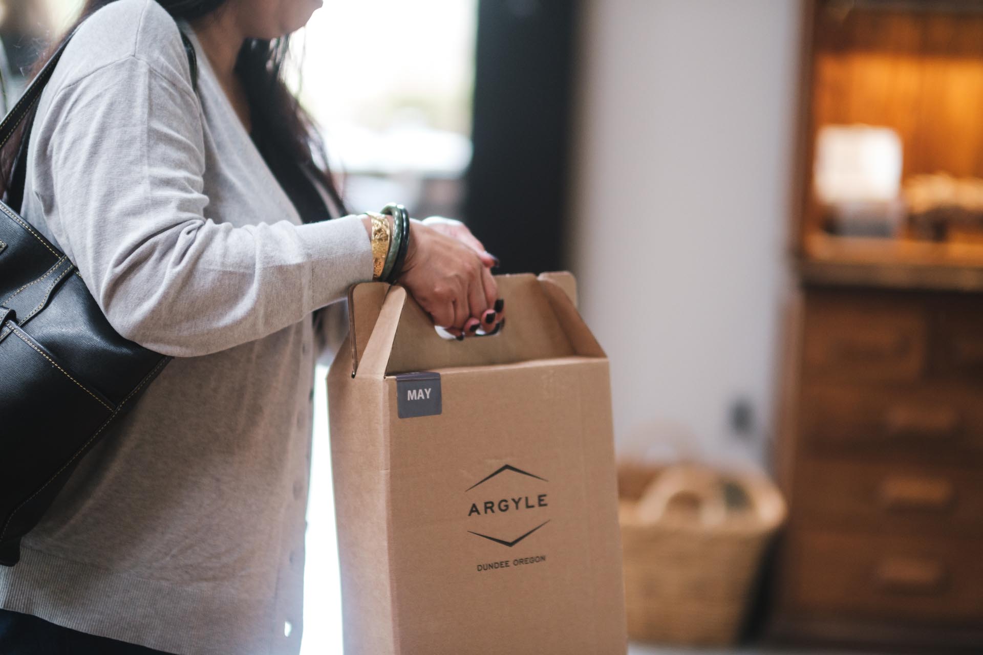 A person wearing a light grey cardigan is holding a brown Wine Club box with Argyle and Dundee Oregon printed on it, along with a small label that reads May. The person has a black handbag over their shoulder. A wooden cabinet is blurred in the background.