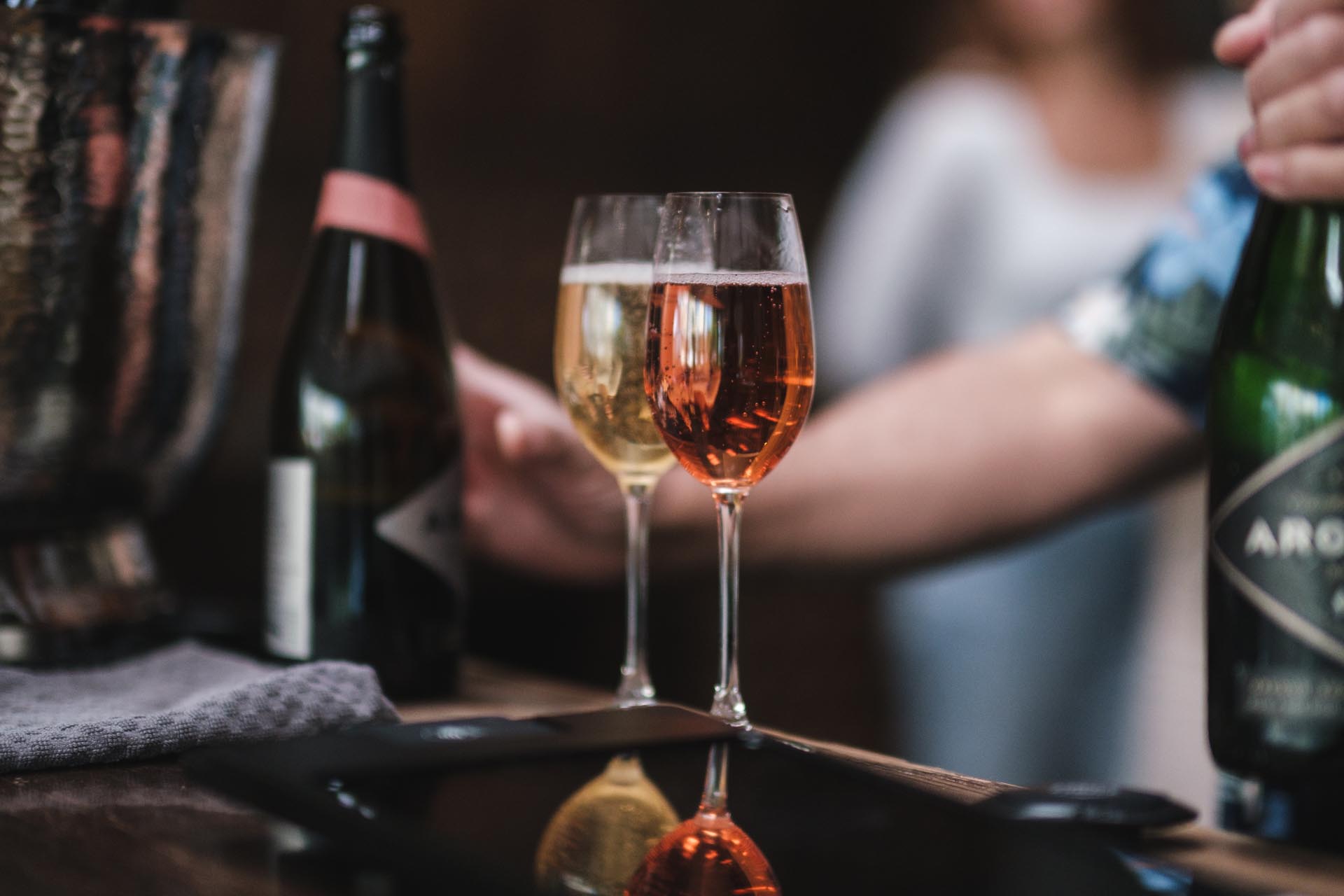 Two wine glasses on a bar counter, one filled with white wine and the other with rosé. A person is holding a bottle and appears to be pouring a drink. Another bottle is partially visible on the counter in the background.