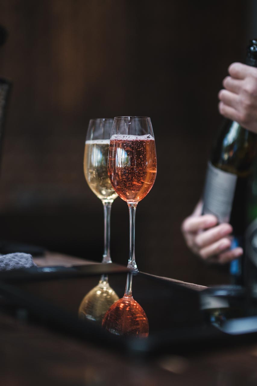 Two glasses of sparkling wine, one with a pale golden liquid and the other with a rosé liquid, are placed on a reflective surface. A person on the right side of the image is holding a wine bottle, partially out of view. The background is dark and blurred.