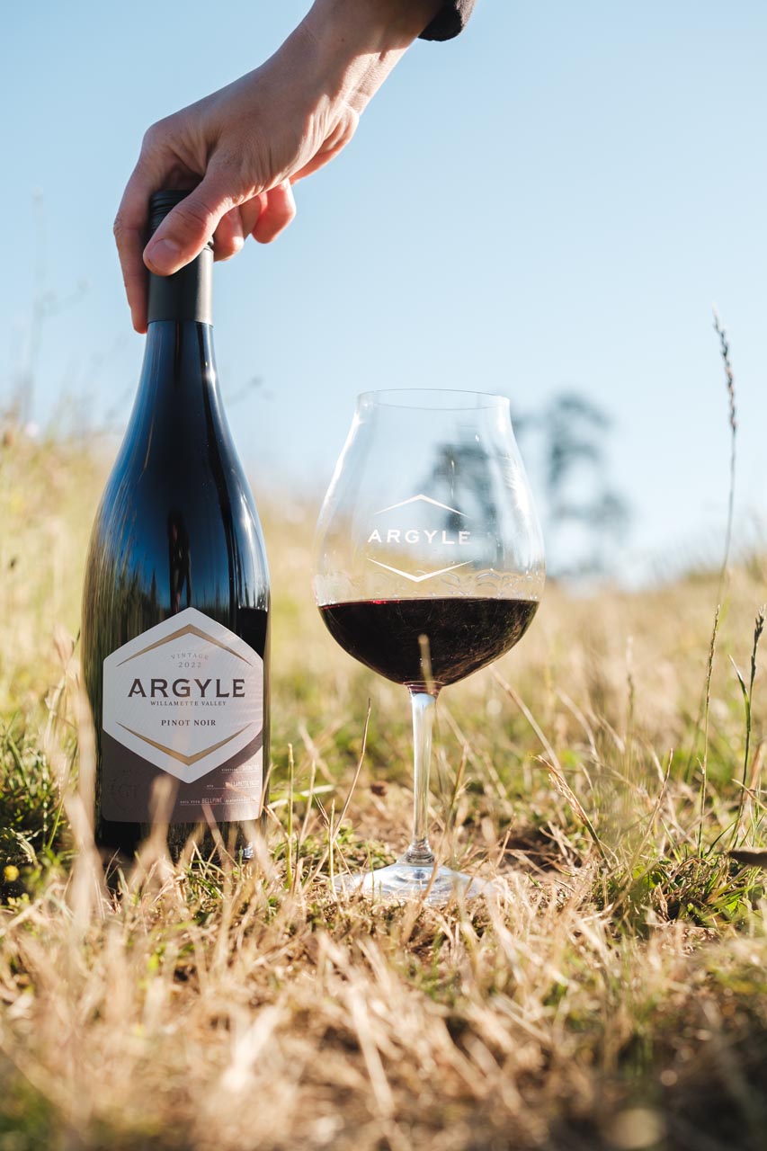 A hand holding the top of a bottle of Argyle Pinot Noir, standing on dry grass in a sunlit field. Beside the bottle is a wine glass filled with red wine, both labeled 