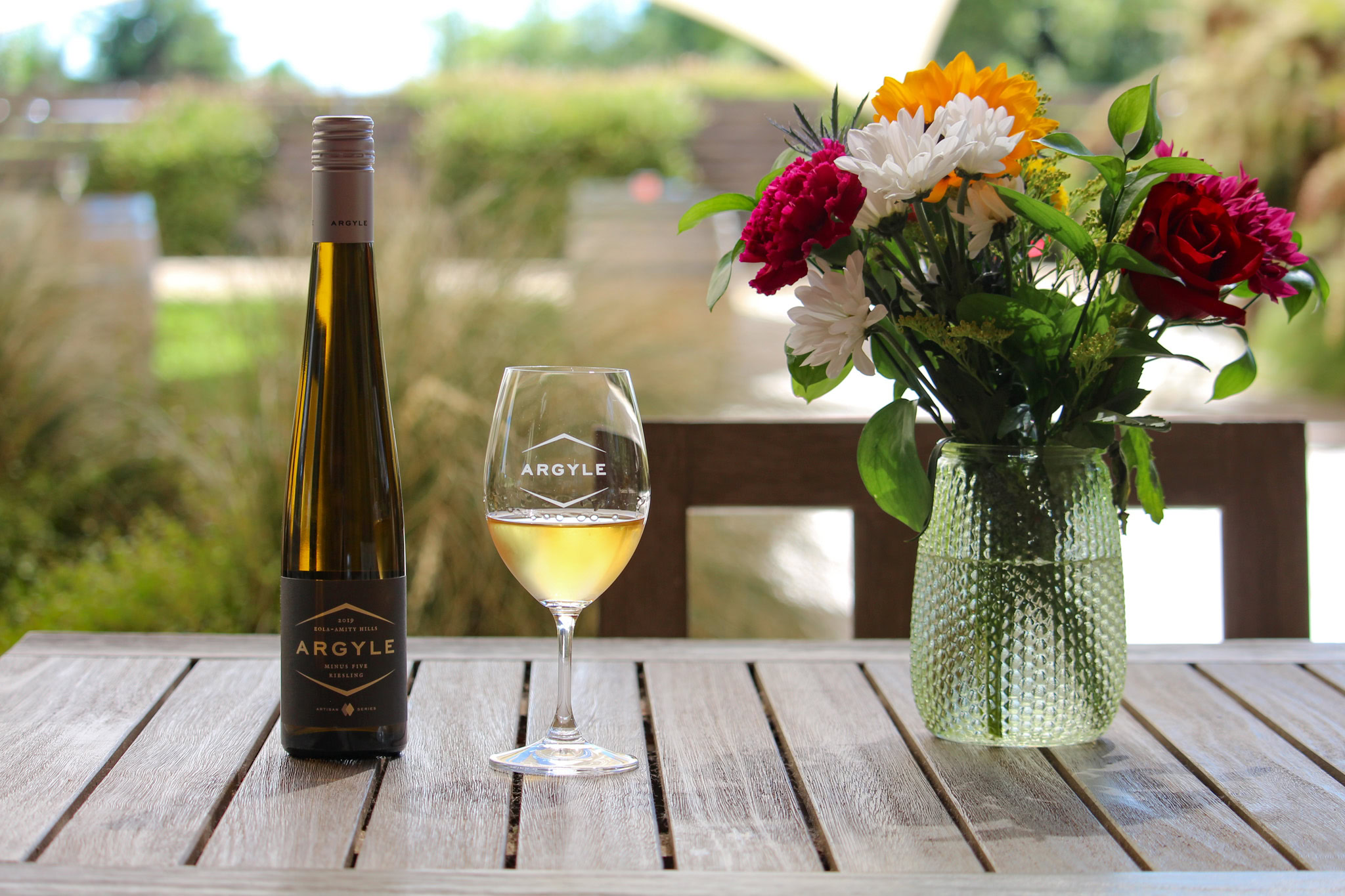 A bottle of Argyle Riesling and a filled wine glass sit on a wooden table. A vase filled with colorful flowers, including red, yellow, and white blossoms, is placed beside the glass. The background features a blurred outdoor setting with greenery and structures.